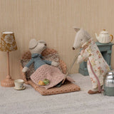 Two toy mice in a cosy room setting with a lamp, tea set, and fireplace.