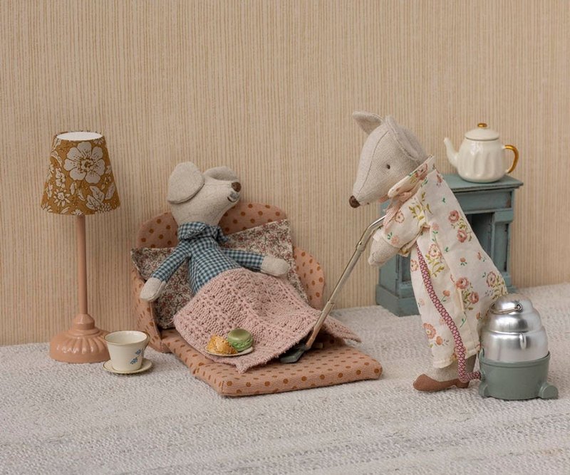 Two toy mice in a cosy room setting with a lamp, tea set, and fireplace.