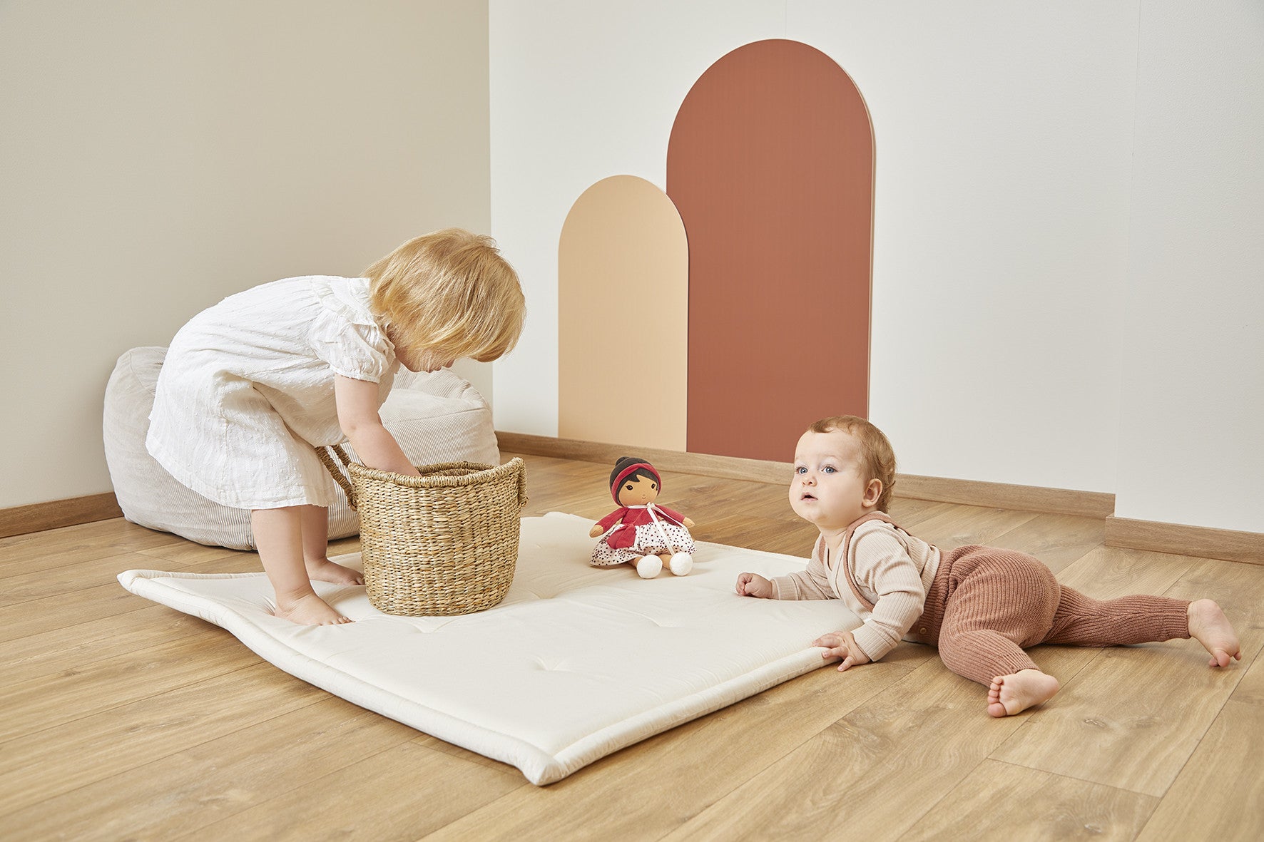 Two children playing with Emilie doll on a white mat wooden floor with a colourful wall in the background.