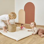 Two children playing with Emilie doll on a white mat wooden floor with a colourful wall in the background.