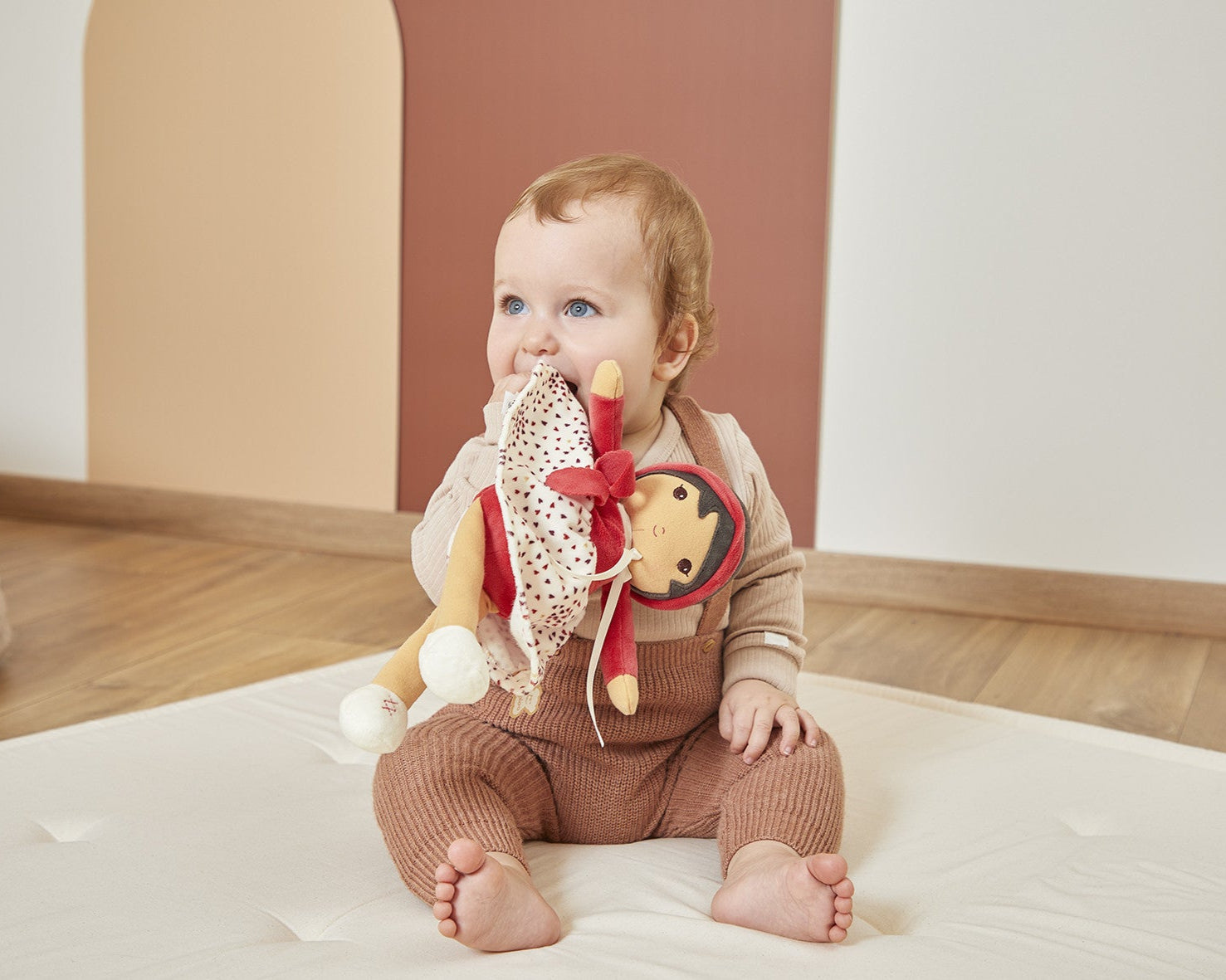 Baby sitting on a white mat with Emilie Doll in a room.