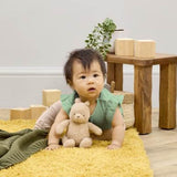 Baby sitting on a yellow rug holding a teddy bear in a room with wooden blocks and a small table.