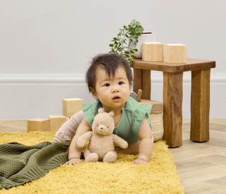 Baby sitting on a yellow rug holding a teddy bear in a room with wooden blocks and a small table.
