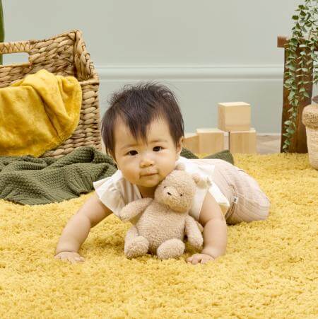 Baby holding a teddy bear on a yellow carpet with a wicker basket and blocks in the background.