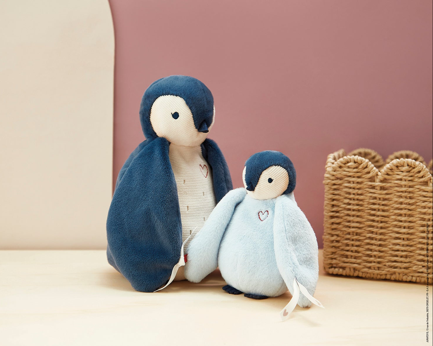 Two toy penguins, one larger and one smaller, sitting on a light surface with a woven basket in the background.