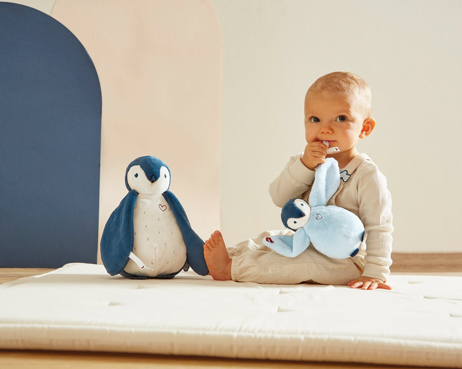 Baby sitting on a bed with two plush penguin toys, one resembling a penguin and the other a rabbit.