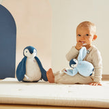 Baby sitting on a bed with two plush penguin toys, one resembling a penguin and the other a rabbit.
